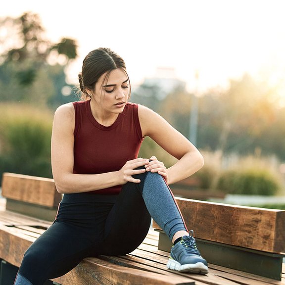 Eine Frau in sportlicher Kleidung sitzt auf einer Parkbank und fasst sich schmerzvoll an ihr Knie.