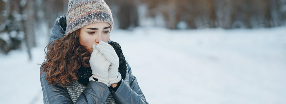 Eine junge Frau steht im Schnee. Sie trägt Winterkleidung und haucht in ihre Handschuhe.