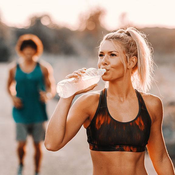 Eine Frau in Sportkleidung trinkt Wasser aus einer Flasche. Im Hintergrund joggt eine Person auf sie zu.