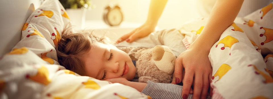 Ein Kind liegt schlafend mit Teddy im Bett. Eine Frau legt schützend die Hände auf seine Arme