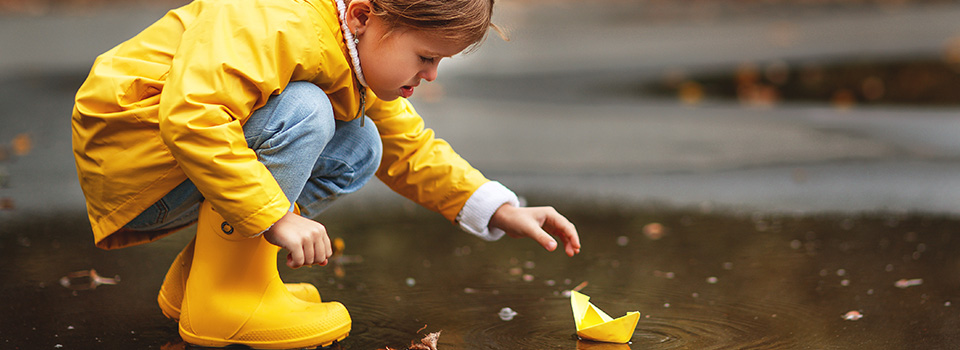 Ein Kind in Regenmantel und Gummistiefel sitzt in der Hocke und greift nach einem Papierboot, das auf  der Oberfläche einer Pfütze treibt.