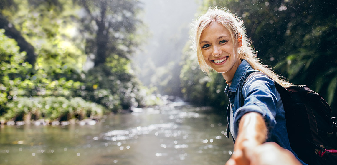 Eine junge Frau wandert an einem Fluss und hält die Hand des Fotografen.
