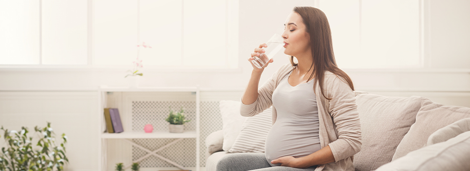 Eine Schwangere sitzt im Schneidersitz auf einem Bett und trinkt Wasser aus einem Wasserglas.